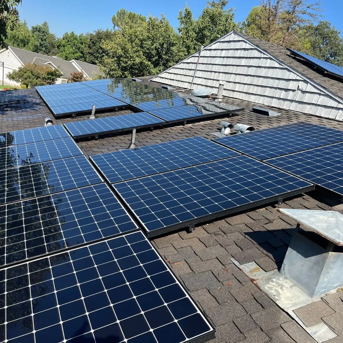 Solar panels on a shingled rooftop under a clear blue sky, surrounded by green trees. Panels glisten in sunlight, conveying sustainable energy.