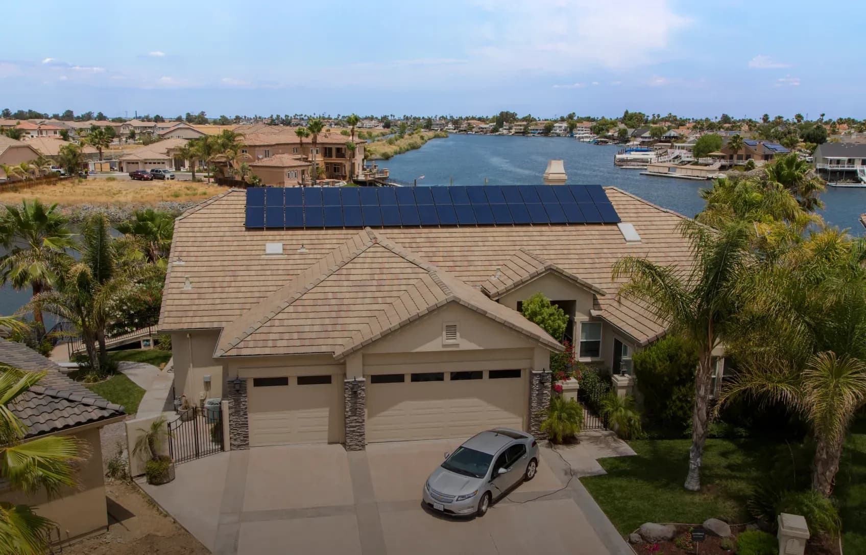 A suburban house with solar panels on the roof sits by a tranquil canal lined with other homes. A car is parked in the driveway, and palm trees frame the scene.