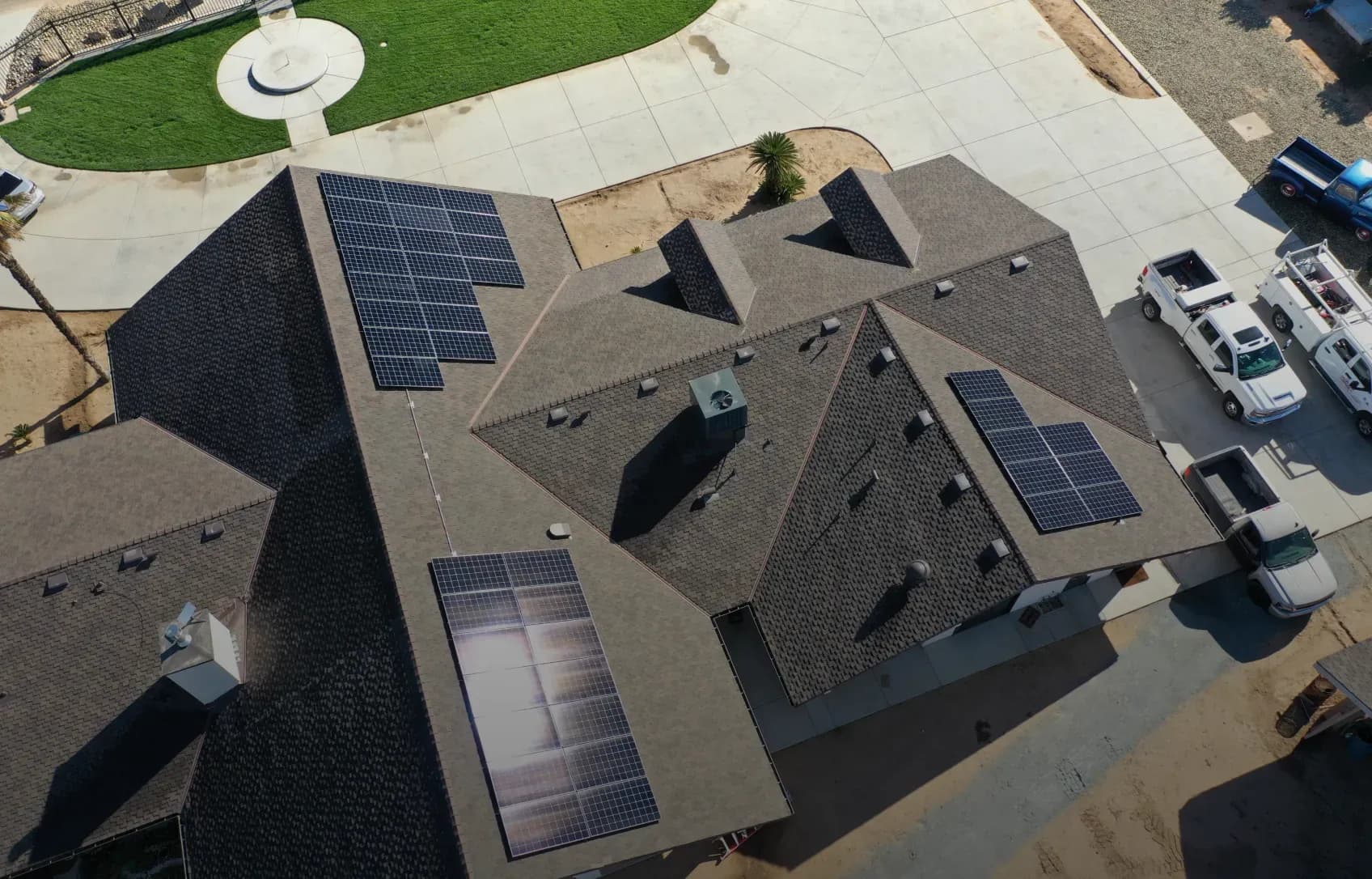 Aerial view of a house with multiple solar panels on its roof. Nearby, several parked trucks are visible on a wide driveway. The scene conveys sustainability.
